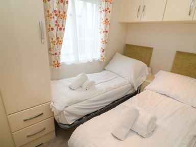 A bedroom with two single beds, beige curtains with floral pattern, and overhead cabinets at The Bobbin, Number 11 in South Lakeland Leisure Village near Carnforth