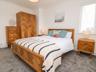 A bedroom with a wooden bed frame white bedding blue pillows wooden wardrobe chest of drawers and bedside table with lamps at Greenbank in Woolacombe