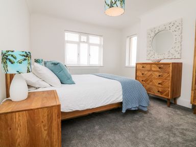 A bedroom with a bed a wooden nightstand with a lamp two windows a wooden dresser and a decorative mirror at Greenbank in Woolacombe