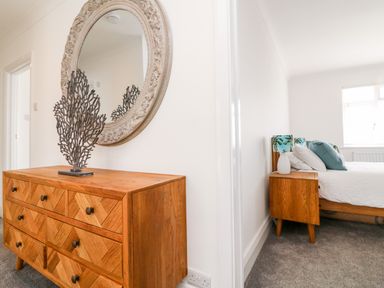 A wooden dresser with a decorative metal sculpture and a round ornate mirror on the wall next to a bedroom with a bed and wooden nightstand with a lamp at Greenbank Woolacombe