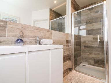 A bathroom with a white sink cabinet a glass soap dispenser a rolled towel a glass shower enclosure and brown tiled walls at Greenbank Woolacombe