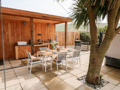 An outdoor dining area with a wooden table and six chairs under a covered wooden structure with a tree nearby at Greenbank in Woolacombe
