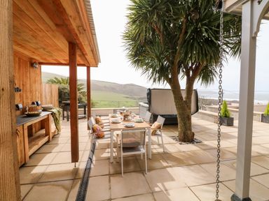 An outdoor patio with a dining table set with plates and glasses near a tree and a wooden shelter with a counter and barbecue at Greenbank Woolacombe