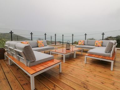An outdoor wooden deck with grey cushioned seating and a wooden table with glass railings at Greenbank in Woolacombe