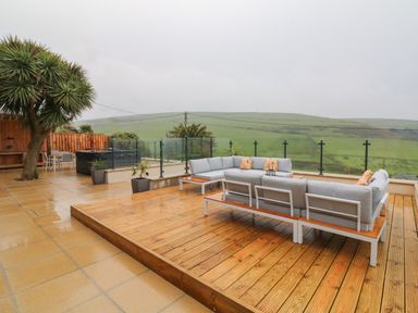 An outdoor patio with a sectional sofa on a wooden deck and glass railing overlooking green hills at Greenbank in Woolacombe
