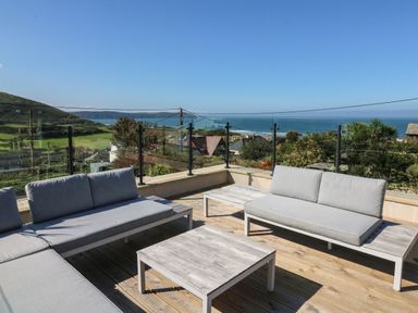 An outdoor terrace with gray cushioned sofas and wooden tables overlooking the sea and hills at Greenbank in Woolacombe