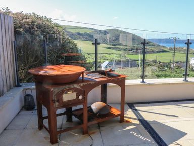 An outdoor patio with a rusty grill and glass railing overlooking green hills and a distant body of water at Greenbank in Woolacombe