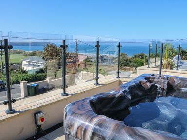 A hot tub on a balcony with glass railing overlooking houses trees and the sea at Greenbank in Woolacombe