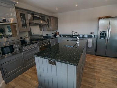 A kitchen with grey cupboards a central island with a sink stainless steel appliances and wooden floor at Nightingales in Bridlington