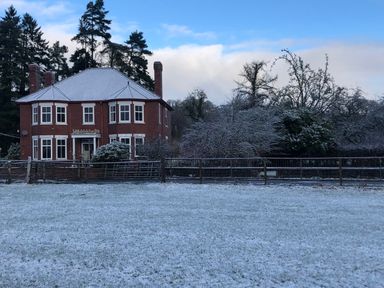 A two-story brick house with a snow-covered roof behind a metal fence and a snowy field with trees in the background at Tirallen in Llangadog