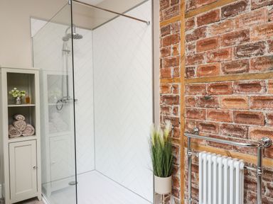 A bathroom with a glass shower enclosure white tiled walls a storage cabinet with towels and a vase next to an exposed brick wall with a radiator