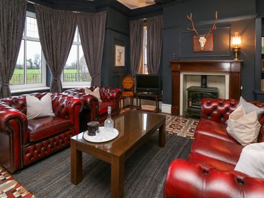 A living room with red leather sofas a wooden coffee table a fireplace and windows with curtains at Tirallen in Llangadog