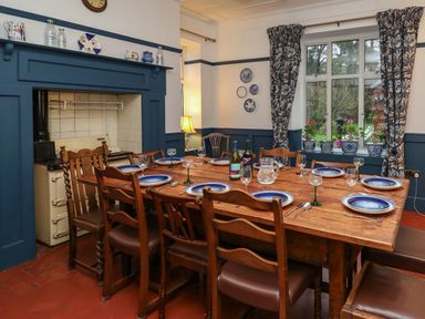 A dining room with a wooden table set with plates and glasses near a window with plants and floral curtains at Tirallen in Llangadog