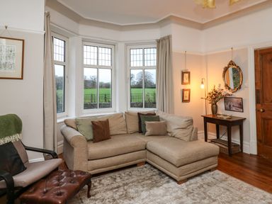 A living room with a beige sofa and cushions a leather footstool beside an armchair a wooden side table with a vase and mirror and windows showing green fields at Tirallen in Llangadog