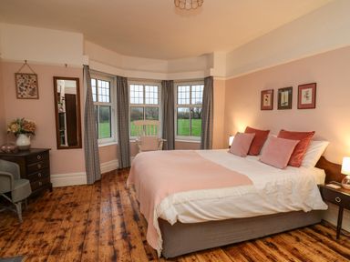 A bedroom with a bed with pillows and a blanket a wooden floor a rocking chair near windows and framed pictures on the wall at Tirallen in Llangadog