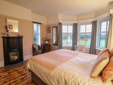 A bedroom with a bed, armchair, small dresser, rocking chair, fireplace, and multiple windows at Tirallen in Llangadog