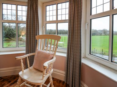 A wooden rocking chair with a cushion by three windows with patterned curtains overlooking a green field at Tirallen in Llangadog