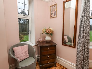 A corner of a room with a gray chair with a pink cushion beside a wooden chest of drawers with flowers in a vase and a hanging mirror at Tirallen in Llangadog