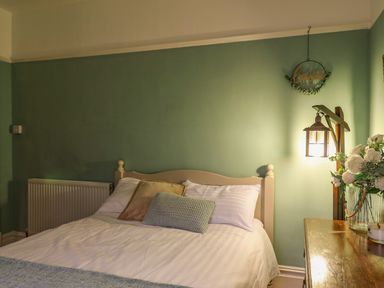 A bedroom with a bed with pillows a wooden bedside table with a lamp and flowers and a wall decoration at Tirallen in Llangadog