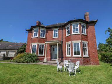 A red brick house with white framed windows and a green lawn with white outdoor chairs and table at Tirallen in Llangadog