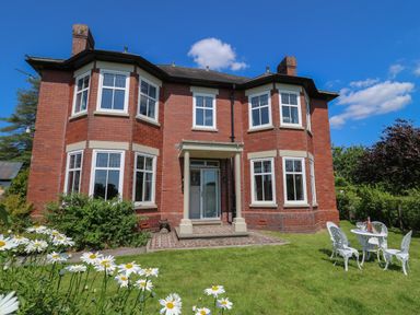 A brick house with white framed windows and a small porch in a garden with daisies and white outdoor table and chairs at Tirallen in Llangadog