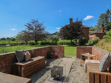 An outdoor seating area with wooden benches and cushions a metal fire pit and a small cart near a green lawn and trees at Tirallen in Llangadog