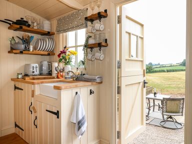 A kitchen corner with shelves of dishes and utensils and an open door leading outside at High Grounds Shepherd's Hut Boylestone near Ashbourne