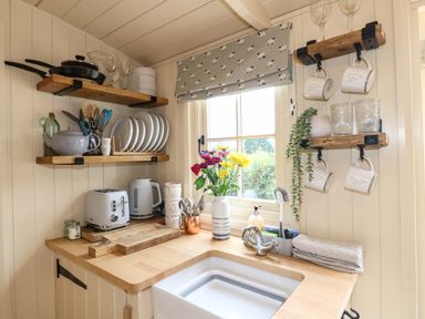 A small kitchen corner with wooden shelves holding plates cups and pans near a window with flowers on the countertop at High Grounds Shepherd's Hut Boylestone near Ashbourne