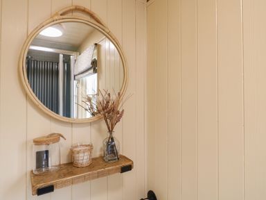 A round mirror above a wooden shelf with a glass jar candle and a vase with dried flowers at High Grounds Shepherd's Hut in Boylestone near Ashbourne