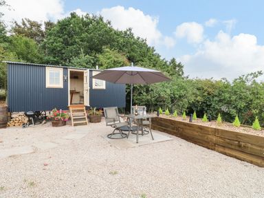 An outdoor patio area with a table and two chairs under an umbrella in front of a black corrugated metal shepherd's hut at High Grounds Shepherd's Hut in Boylestone near Ashbourne