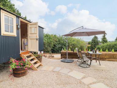 An outdoor patio area with gravel and stepping stones outside a small building with an open door flowers in wooden barrels and a table with chairs under an umbrella at High Grounds Shepherd's Hut in Boylestone near Ashbourne