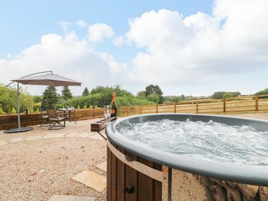 An outdoor area with a hot tub bubbling a table with two chairs and an umbrella overlooking fields at High Grounds Shepherd's Hut Boylestone near Ashbourne