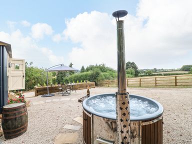 A hot tub on a gravel patio with a table and chairs under an umbrella and a wooden fence in the background at High Grounds Shepherd's Hut in Boylestone near Ashbourne