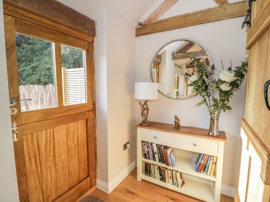 A hallway with a wooden door a white console table with books flowers and a lamp below a round mirror at The Old Piggery in Defford