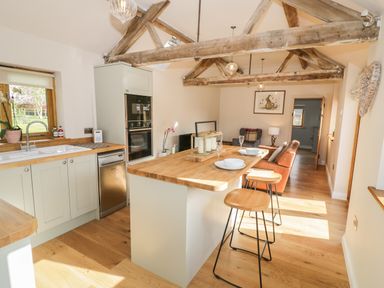 An open kitchen and living area with wooden beams on the ceiling a kitchen island with stools and a leather sofa at The Old Piggery in Defford