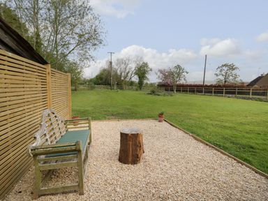 An outdoor seating area with a wooden bench and tree stump table on gravel next to a grassy lawn at The Old Piggery in Defford