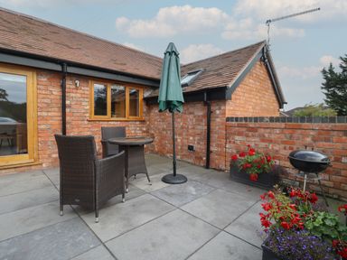 An outdoor patio with wicker chairs a round table a closed umbrella flower pots and a barbecue at The Old Piggery Defford