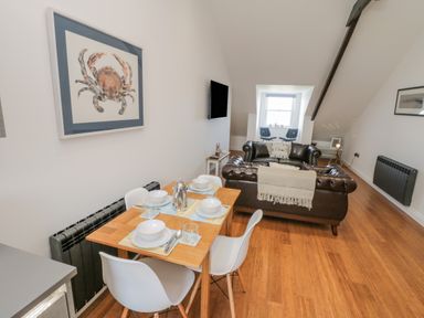A dining area with a wooden table and white chairs next to a living room with brown leather sofas and a wall-mounted television at Foxton Penthouse in Whitby