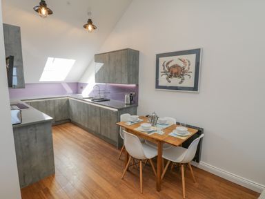 A kitchen with gray cabinets and a wooden floor and a dining table set with white chairs and a crab picture on the wall at Foxton Penthouse in Whitby