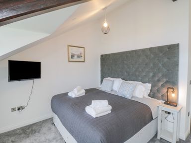 A bedroom with a bed with gray quilted headboard and bedding a small bedside table with lamp and a wall-mounted television at Foxton Penthouse in Whitby