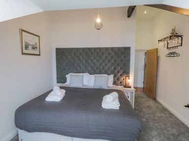 A bedroom with a large bed with gray quilt and pillows with a tufted headboard a side table with lamp a wooden door and wall shelf with hangers at Foxton Penthouse in Whitby