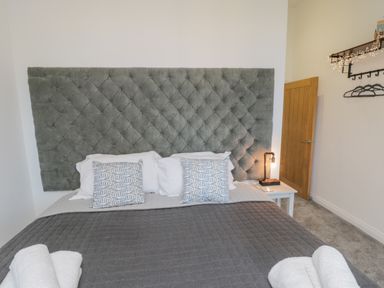 A bedroom with a large upholstered headboard a bed with pillows a bedside table with a lamp and folded towels at Foxton Penthouse in Whitby