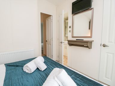 A bedroom with a bed covered in a teal blanket and white towels on top a wall-mounted mirror and television are opposite two open doors showing a bathroom and another room at Number 10 in Warton