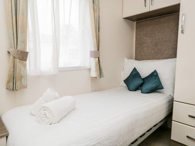 A single bed with white linens two teal pillows and rolled white towels by a window with curtains at Number 10 in Warton