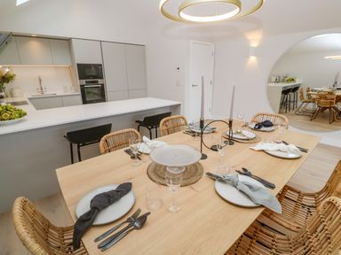 A dining table set with plates glasses and cutlery in a kitchen with rattan chairs and a large round mirror at Porth Beach in Porth near St Columb Minor