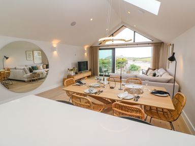 A dining table with place settings and rattan chairs in an open living room with a large round wall mirror at Porth Beach in Porth near St Columb Minor