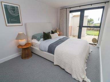 A bedroom with a bed with pillows and blankets a wooden side table with a lamp a framed artwork on the wall and a glass door to the outside at Porth Beach in Porth near St Columb Minor