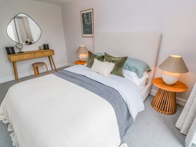 A bedroom with a bed a wooden bedside table with a lamp a wooden dressing table with a stool and a wall mirror at Porth Beach in Porth near St Columb Minor