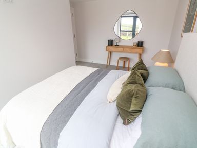 A bedroom with a bed with green and beige pillows a bedside lamp a wooden dressing table with a mirror and stool at Porth Beach in Porth near St Columb Minor