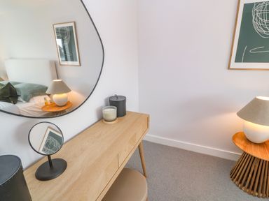 A bedroom corner with a wooden desk, round mirror, lamp on a wooden side table, and a framed abstract print on the wall at Porth Beach in Porth near St Columb Minor
